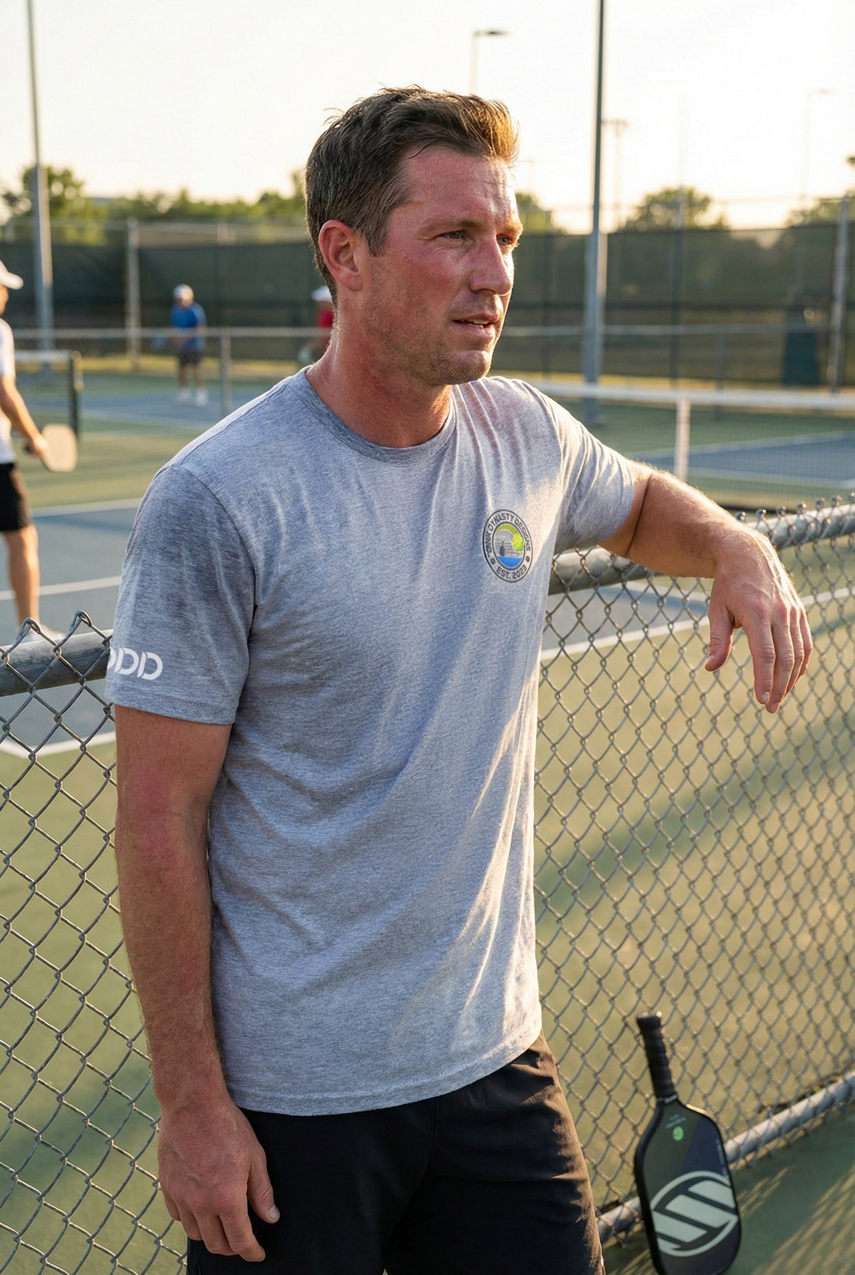 Man standing on a tennis court with a pickleball paddle, wearing a gray t-shirt.