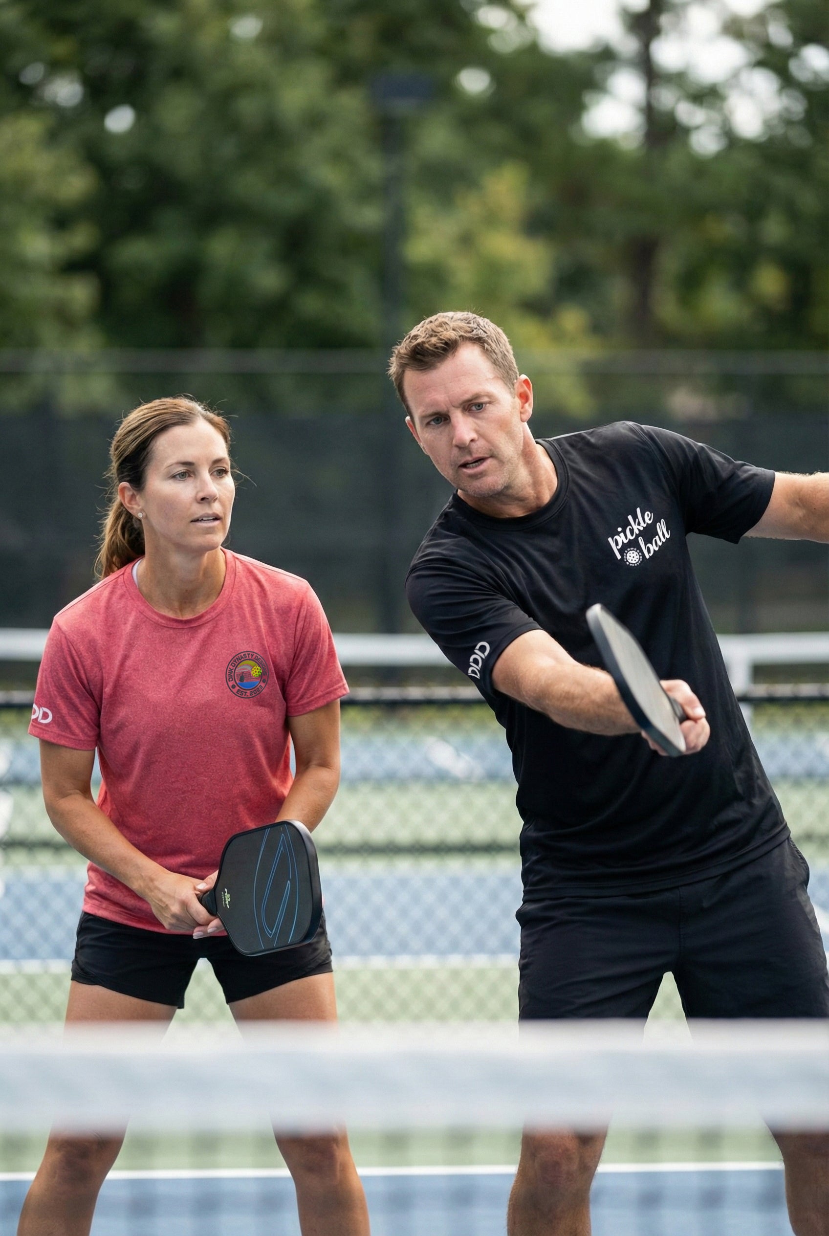 Two people playing pickleball on a court with trees in the background